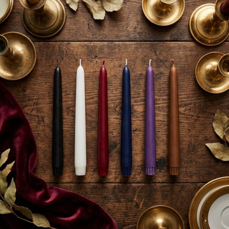 Row of unlit black, white, red, blue, purple and brown taper candles, each a different protection candle color, on a wooden table ringed by brass candlesticks