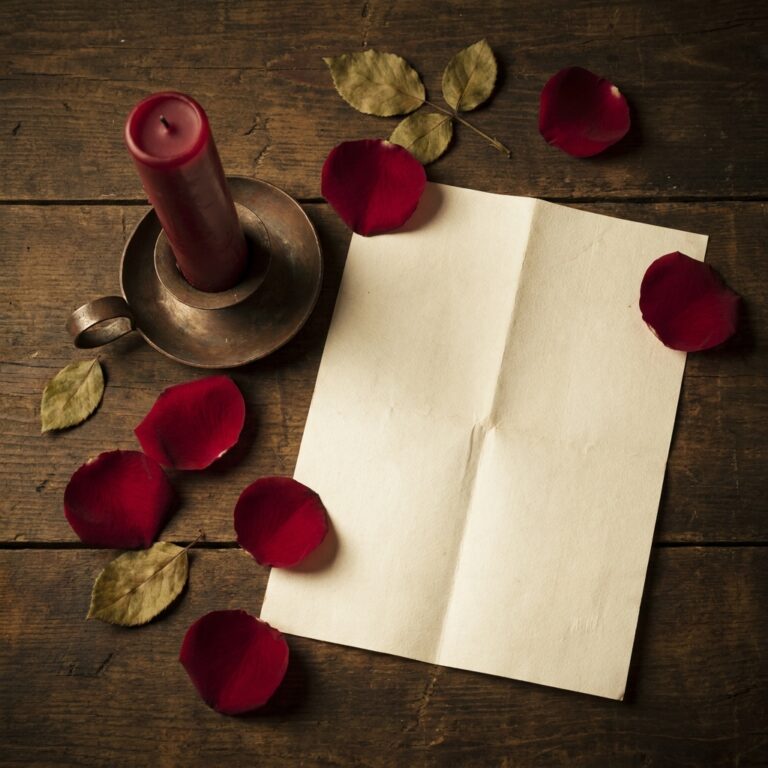 A blank creased paper, an unlit red candle and scattered rose petals on dark wood, the first materials of a traditional obsession spell