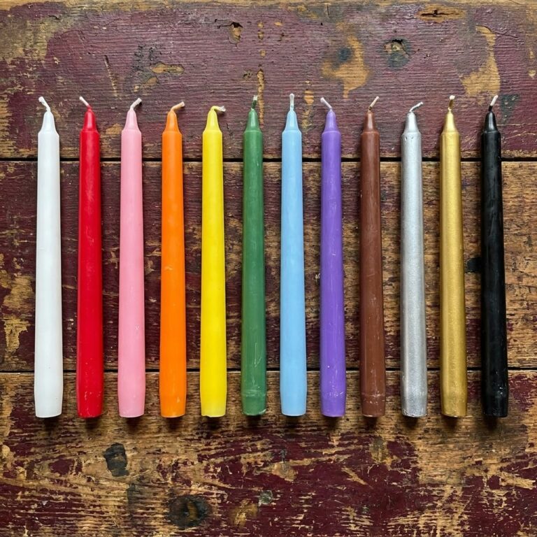 Twelve unlit taper candles laid side by side on weathered wood, from white through red, green, purple, silver and gold to black, showing the range of candle colors used in spells