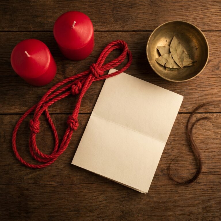 Flat-lay for a love binding spell: two red candles, a red cord tied in four knots, a blank folded paper, a strand of dark hair and a brass bowl of bay leaves on dark wood